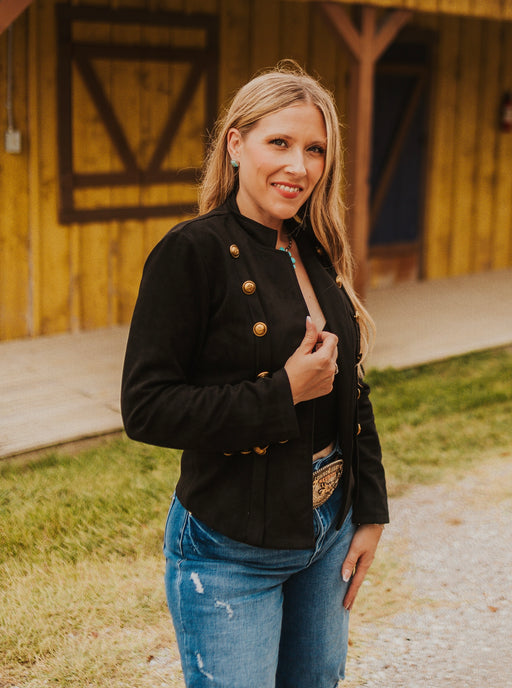 Woman wearing a black jacket with gold buttons and blue jeans standing in front of a wooden building.