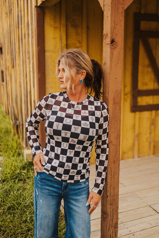 Woman wearing a black and white checkered shirt and blue jeans standing against a wooden structure.