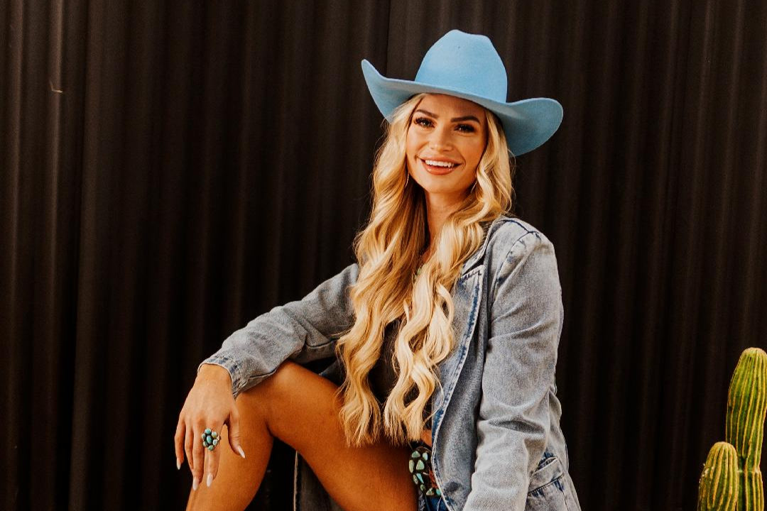 Woman wearing a blue cowboy hat and gray jacket sitting against a dark background with a cactus plant.
